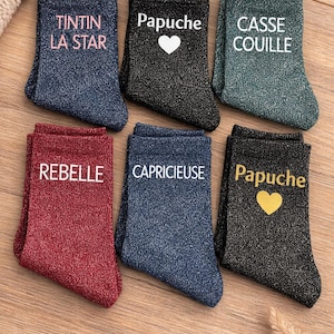 Peut inclure: Six paires de chaussettes scintillantes de différentes couleurs, chacune avec un mot ou une phrase différente en blanc ou en or. Les chaussettes sont bleues, noires, vertes, rouges et marine. Les mots incluent "TINTIN LA STAR", "Papuche", "CASSE COUILLE", "REBELLE", "CAPRICIEUSE", et "Papuche".