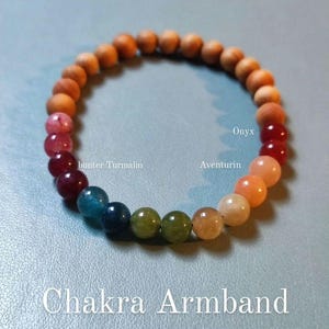 Könnte beinhalten: Ein Chakra-Armband aus natürlichen Steinen, darunter Onyx, Aventurin und rosa Turmalin. Das Armband ist auf einer Holzschnur aufgefädelt und hat ein Regenbogen-Farbschema. Der Text "Chakra Armband" steht unter dem Armband. Der Text "wie immer: kann individuell nach deinem Wunsch zusammengestellt werden" steht unter dem Text "Chakra Armband".