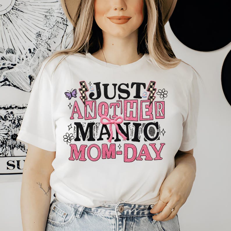 May include: White t-shirt with the text "JUST ANOTHER MANIC MOM-DAY" in pink and black lettering. The design includes butterflies, flowers, and a pink bow. The shirt is short-sleeved.