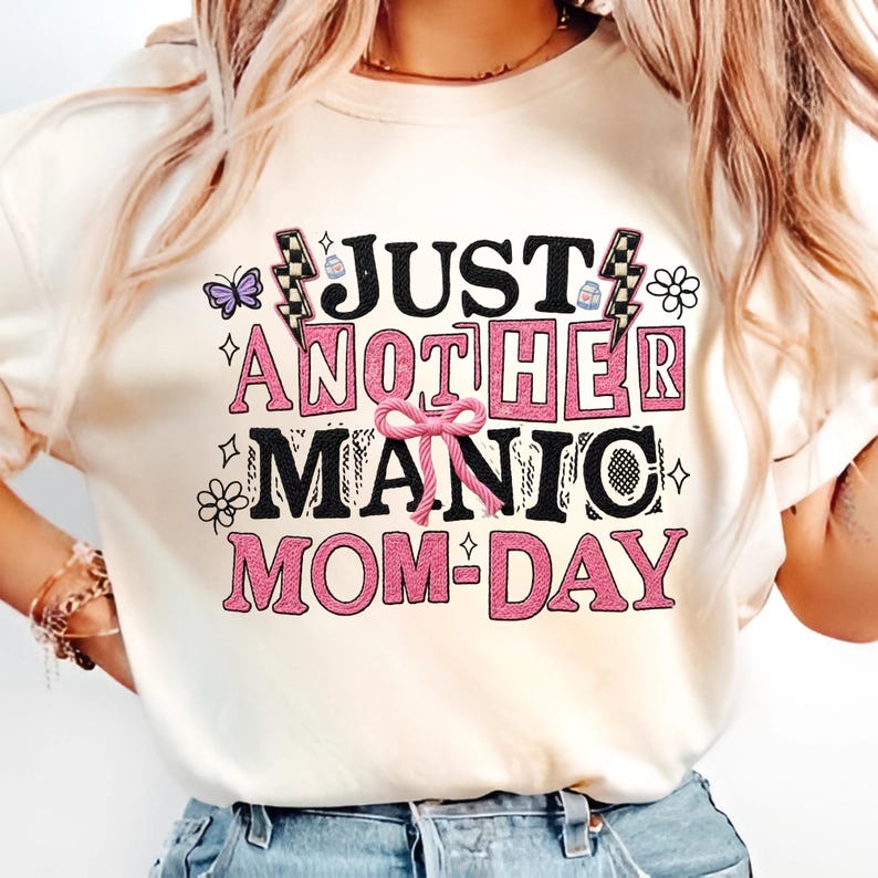 May include: Cream-colored t-shirt with the text "JUST ANOTHER MANIC MOM-DAY" in black and pink block letters. The design includes a pink bow, butterflies, flowers, and lightning bolts. A casual, everyday top.