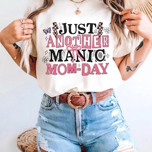 May include: Cream-colored t-shirt with the text "JUST ANOTHER MANIC MOM-DAY" in pink and black. The design includes butterflies, flowers, and lightning bolts. The shirt is paired with blue denim shorts and a brown belt.