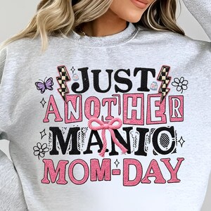 May include: A light gray sweatshirt with the text "JUST ANOTHER MANIC MOM-DAY" in black and pink. The design includes a butterfly, lightning bolts, and floral accents.