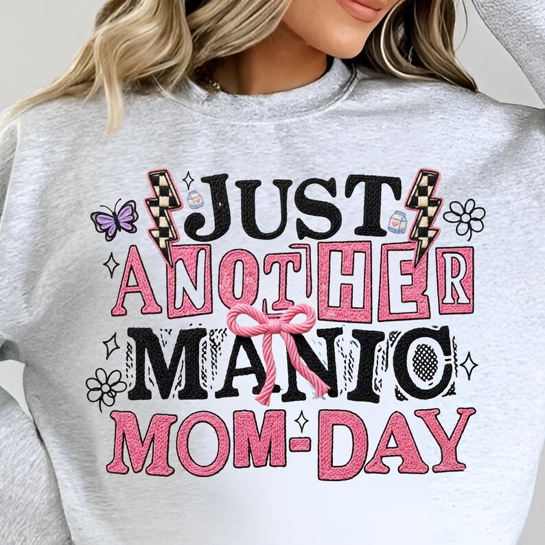 May include: A light gray sweatshirt with the text "JUST ANOTHER MANIC MOM-DAY" in black and pink. The design includes a butterfly, lightning bolts, and floral accents.