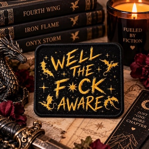 Puede incluir: Un parche rectangular negro bordado con hilo dorado que forma el texto WELL THE F*CK AWARE, acentuado con pequeñas siluetas de dragones y estrellas bordadas. El parche tiene un borde cosido y una base de tela oscura y texturizada.