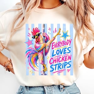 May include: A cream-colored t-shirt featuring a colorful graphic of a rooster wearing sunglasses and dancing on a pole. The design includes blue vertical stripes, pink and yellow stars, and the text "EVERYBODY LOVES CHICKEN STRIPS" in blue and pink.