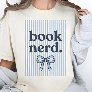 May include: Cream-colored t-shirt with the text "book nerd." in navy blue, set against a light blue and white striped background. A light blue bow graphic is below the text. The shirt has a round neckline and short sleeves.