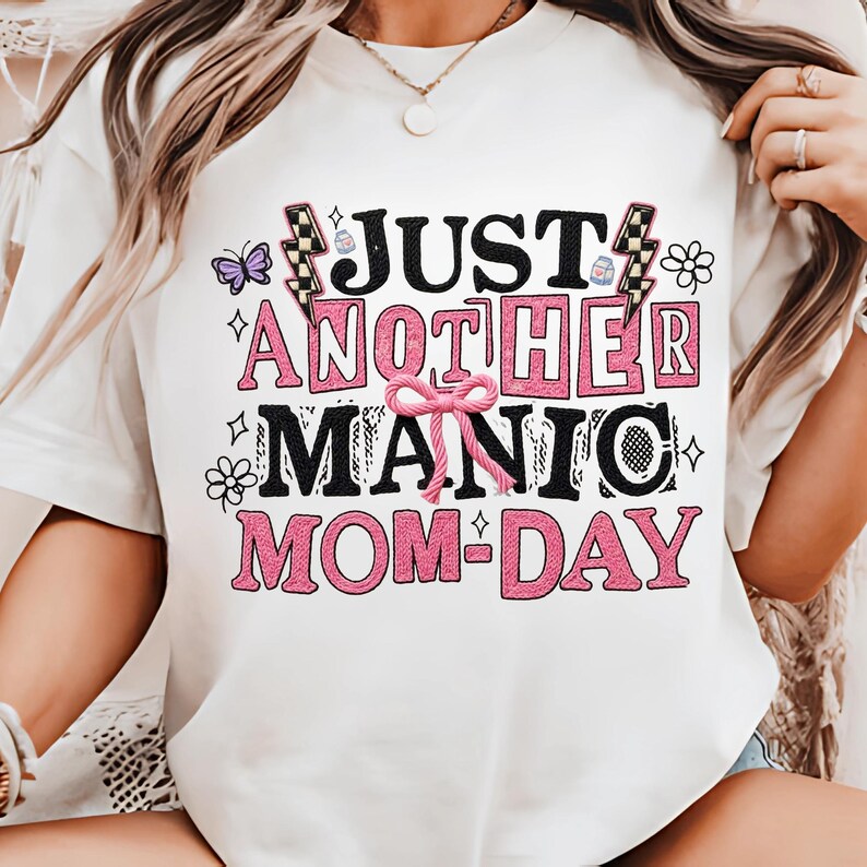 May include: White t-shirt with the text "JUST ANOTHER MANIC MOM-DAY" in black and pink. The design includes a pink bow, lightning bolts, butterflies, and flowers. The shirt is a casual, everyday wear item.