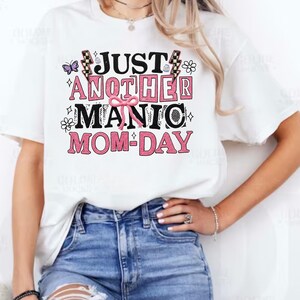 May include: White t-shirt with the text "JUST ANOTHER MANIC MOM-DAY" in black and pink. The design includes a butterfly, flowers, and lightning bolts. The shirt is paired with blue jeans.