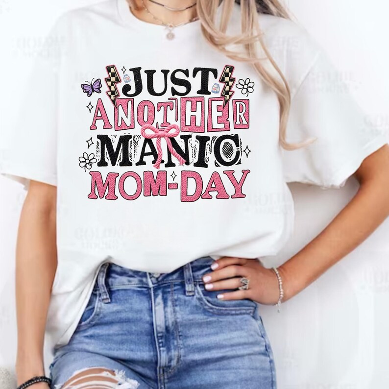 May include: White t-shirt with the text "JUST ANOTHER MANIC MOM-DAY" in black and pink. The design includes a butterfly, flowers, and lightning bolts. The shirt is paired with blue jeans.