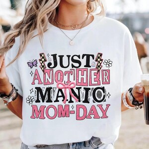 May include: White t-shirt with the text "JUST ANOTHER MANIC MOM-DAY" in black and pink block letters. The design includes a butterfly, flowers, and lightning bolts. The shirt is a casual, everyday wear item.