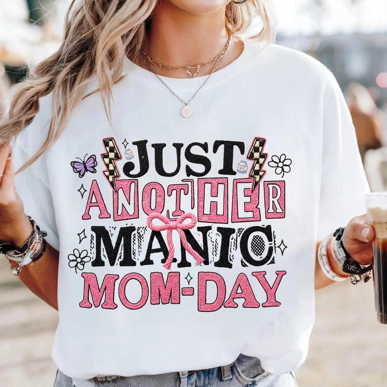 May include: White t-shirt with the text "JUST ANOTHER MANIC MOM-DAY" in black and pink block letters. The design includes a butterfly, flowers, and lightning bolts. The shirt is a casual, everyday wear item.