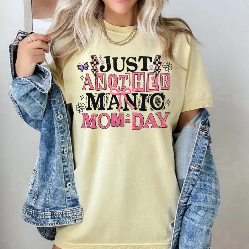 May include: A light yellow t-shirt with the text "JUST ANOTHER MANIC MOM-DAY" in black and pink. The shirt has a relaxed fit and is paired with a blue denim jacket.