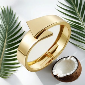 May include: A gold bangle bracelet with a unique, angular design. The bracelet is open and has a hinge closure.
