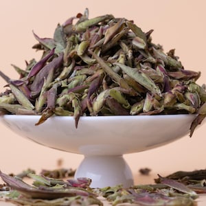 May include: A white ceramic bowl filled with loose leaf tea. The tea is a blend of green and purple leaves.