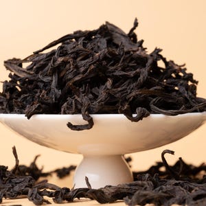 May include: A white ceramic dish filled with loose black tea leaves. The dish is on a white pedestal and there are loose tea leaves scattered around the base of the pedestal.