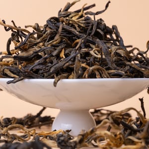 May include: A white ceramic bowl filled with loose leaf black tea. The tea leaves are a mix of dark brown and golden brown colors. The bowl is on a white pedestal.