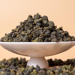 May include: A white ceramic dish filled with loose green tea leaves. The tea leaves are arranged in a mound in the center of the dish. The dish is sitting on a table with more tea leaves scattered around it.