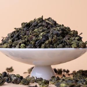 May include: A white, pedestal serving dish overflowing with dried green tea leaves. The tea leaves are a mix of dark green and brown, with some scattered around the base of the dish. The background is a solid, light peach color.