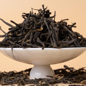 May include: A white ceramic dish filled with loose leaf black tea. The tea leaves are dark brown and have a slightly curled shape.