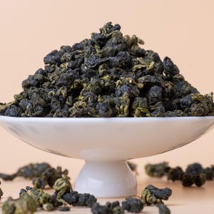 May include: A white, pedestal serving dish overflowing with dark green, tightly rolled tea leaves. Some leaves are scattered around the base of the dish. The background is a solid, light peach color.