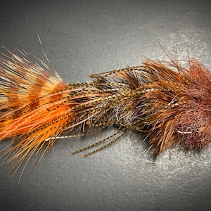 Peut inclure: Mouche de pêche artisanale aux couleurs vives. Elle présente un mélange de plumes orange, marron et beige, créant un aspect texturé. La mouche est conçue pour attirer les poissons et est un choix populaire pour les amateurs de pêche à la mouche.