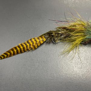 Puede incluir: Una mosca de pesca hecha a mano con un anzuelo. La mosca presenta una cola de plumas a rayas amarillas y negras, que se transforma en un cuerpo de plumas verdes, marrones y amarillas. Diseñada para la pesca.