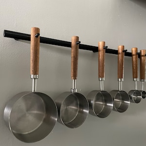 Puede incluir: Un juego de ocho tazas medidoras de acero inoxidable con mangos de madera colgadas de un estante de metal negro contra una pared blanca.