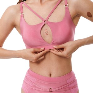 May include: A pink lingerie set comprising a bralette with a keyhole and crisscross straps, and high-waisted briefs. The bralette has gold-coloured hardware. The text "Think Love" is visible on the bralette. The set is made of a soft, smooth fabric.