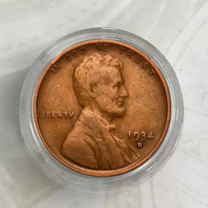May include: A copper-colored 1934 penny coin with the words "In God We Trust" and "Liberty" inscribed on it. The coin features a profile of Abraham Lincoln on one side.