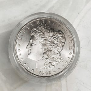 Op de afbeelding: Een zilveren Morgan dollar-munt met het jaar 1892 gegraveerd aan de onderkant. De munt toont een profiel van een vrouw met golvend haar en de inscriptie "E PLURIBUS UNUM" rond de rand.