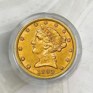 May include: A gold-coloured coin featuring a portrait of a woman wearing a hat with the word "LIBERTY" inscribed on it. The coin has a date of "1893" and a border of stars.