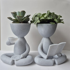 May include: Two grey ceramic planters shaped like people, one holding a cup and saucer, the other holding a book, both with green plants growing out of their heads.