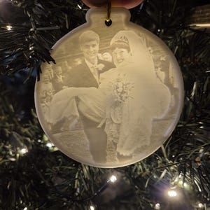 May include: A white ceramic Christmas bauble featuring a photograph of a bride and groom. The bride is wearing a white dress and veil, and the groom is wearing a suit. The bauble is hanging on a Christmas tree.