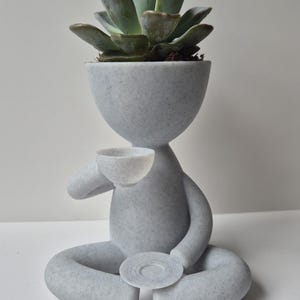 May include: A grey ceramic planter shaped like a person sitting in a meditative pose, holding a small cup in one hand and a saucer in the other. The planter has a textured surface and is topped with a green succulent plant.