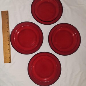 May include: Four circular red glass plates arranged in a diamond pattern. A wooden ruler is positioned vertically on the left side, showing the plates have a diameter of approximately 6 inches. The plates feature a smooth, glossy finish.