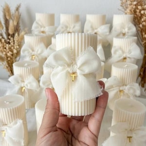 May include: A cylindrical, cream-colored candle with a vertical ribbed texture. The candle is adorned with a frayed-edge, off-white fabric bow tied around the center, featuring a small, gold-toned cross charm attached to the knot of the bow.