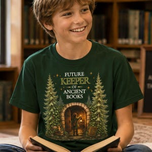 May include: A dark green t-shirt featuring a graphic print of a stone archway flanked by two tall pine trees. The text reads "FUTURE KEEPER OF ANCIENT BOOKS" above an illustration of a person holding a lantern in front of the open stone doorway.