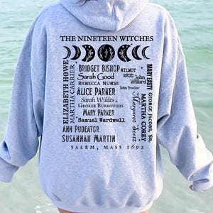 Op de afbeelding: Een asgrijze hoodie met een zwarte opdruk op de achterkant. Het ontwerp bevat maanfasen, de tekst THE NINETEEN WITCHES, een lijst met namen waaronder Bridget Bishop en Sarah Good, en de tekst SALEM, MASS 1692 onderaan het ontwerp.