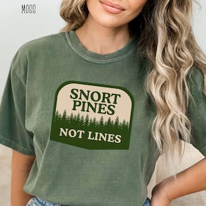 May include: A moss green t-shirt with the text "SNORT PINES NOT LINES" in a white and dark green design. The design features a forest silhouette. The shirt is a short-sleeved, casual style.