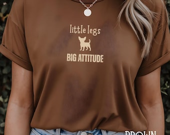 May include: Brown t-shirt with the text "little legs BIG ATTITUDE" and a dog silhouette. The word "BROWN" is printed at the bottom of the shirt. The shirt has rolled-up sleeves and is worn with blue jeans.