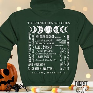 Op de afbeelding: Een bosgroene hoodie met rits en een witte afbeelding op de achterkant. Het ontwerp bevat maanfasen, de tekst THE NINETEEN WITCHES, een lijst met namen waaronder Bridget Bishop en Sarah Good, en de tekst SALEM, MASS 1692 met kleine sterren.