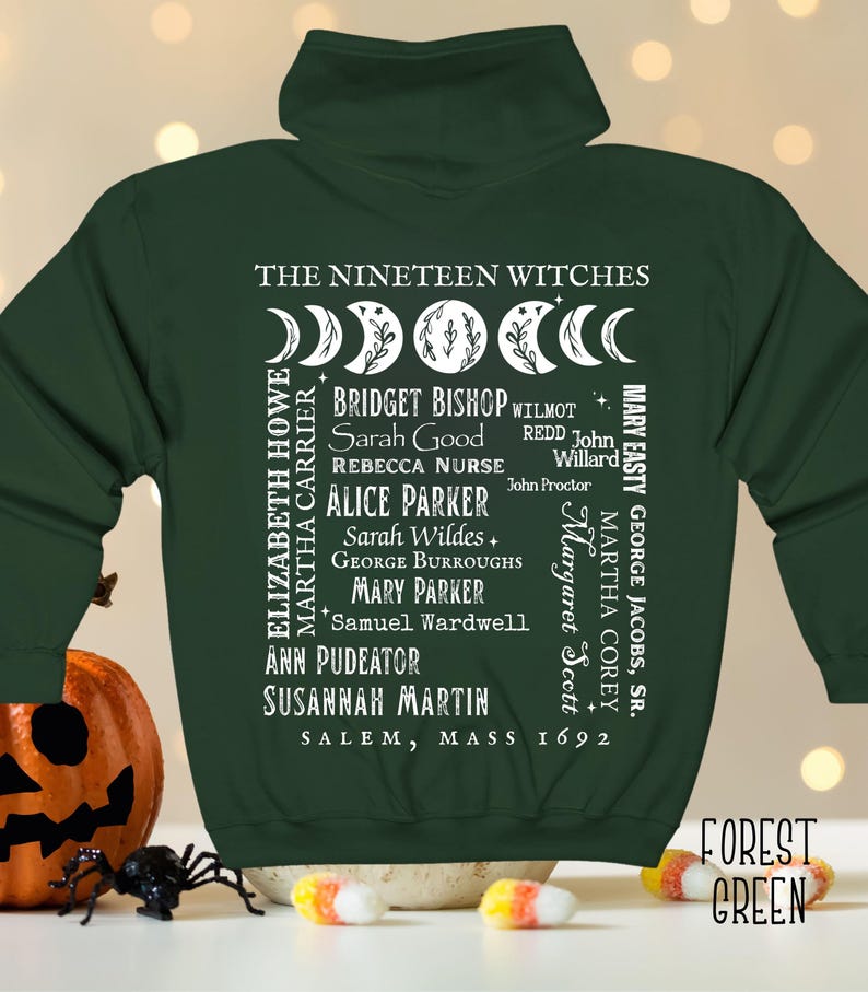 Op de afbeelding: Een bosgroene hoodie met rits en een witte afbeelding op de achterkant. Het ontwerp bevat maanfasen, de tekst THE NINETEEN WITCHES, een lijst met namen waaronder Bridget Bishop en Sarah Good, en de tekst SALEM, MASS 1692 met kleine sterren.