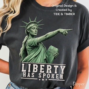 May include: A charcoal gray t-shirt featuring a green graphic of the Statue of Liberty pointing forward while holding a tablet that reads "DON'T YOU POINT AT ME". Below the graphic, the text reads "LIBERTY HAS SPOKEN" with three stars.