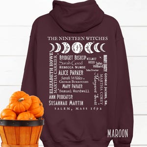 Op de afbeelding: Een kastanjebruine hoodie met een witte afbeelding op de achterkant. De tekst luidt THE NINETEEN WITCHES, SALEM, MASS 1692, en bevat namen zoals Bridget Bishop en Sarah Good, vergezeld van een maanfase-ontwerp met florale details.