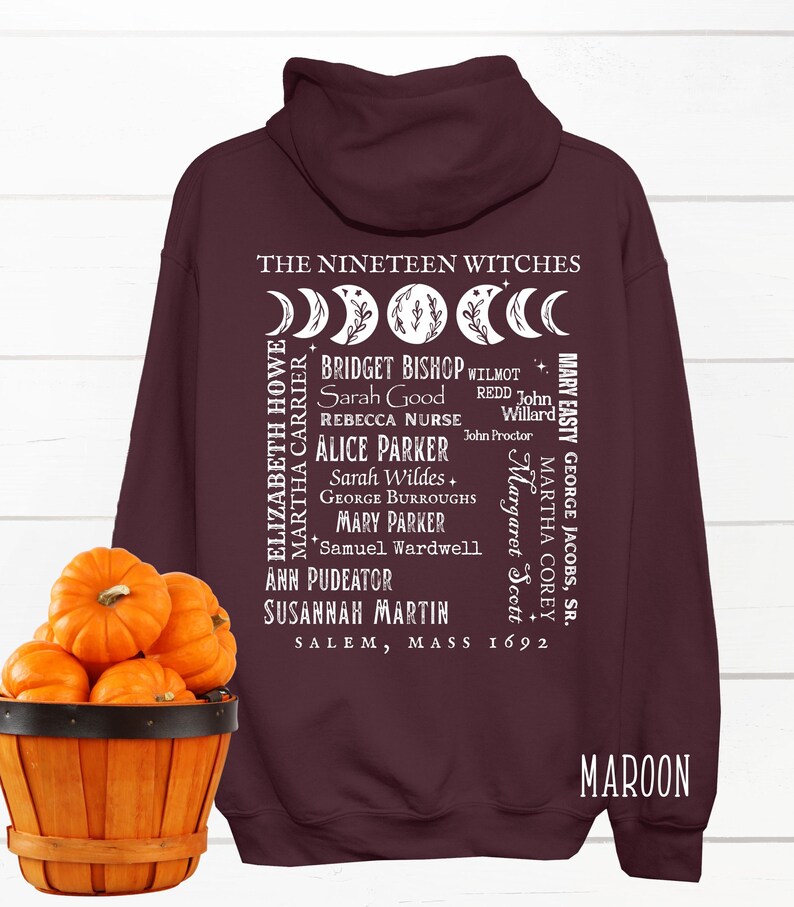 Op de afbeelding: Een kastanjebruine hoodie met een witte afbeelding op de achterkant. De tekst luidt THE NINETEEN WITCHES, SALEM, MASS 1692, en bevat namen zoals Bridget Bishop en Sarah Good, vergezeld van een maanfase-ontwerp met florale details.