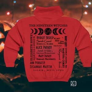 Op de afbeelding: Een rode hoodie met zwarte tekst en afbeeldingen op de achterkant. Het ontwerp bevat de woorden THE NINETEEN WITCHES, een maanfasencyclus, een lijst met namen waaronder Bridget Bishop en Sarah Good, en SALEM, MASS 1692.