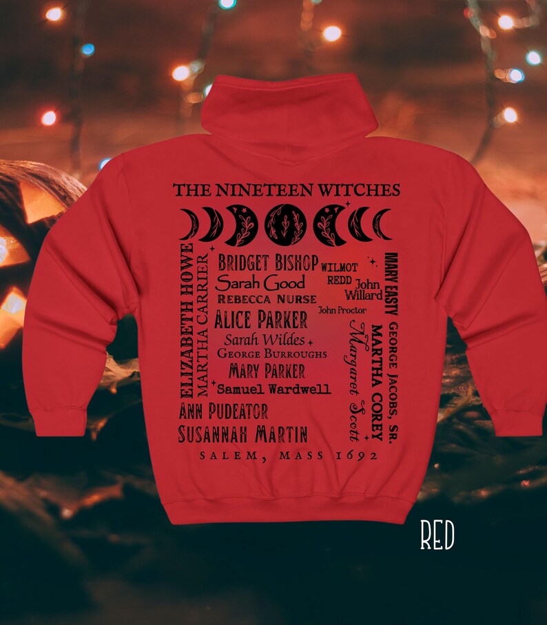 Op de afbeelding: Een rode hoodie met zwarte tekst en afbeeldingen op de achterkant. Het ontwerp bevat de woorden THE NINETEEN WITCHES, een maanfasencyclus, een lijst met namen waaronder Bridget Bishop en Sarah Good, en SALEM, MASS 1692.
