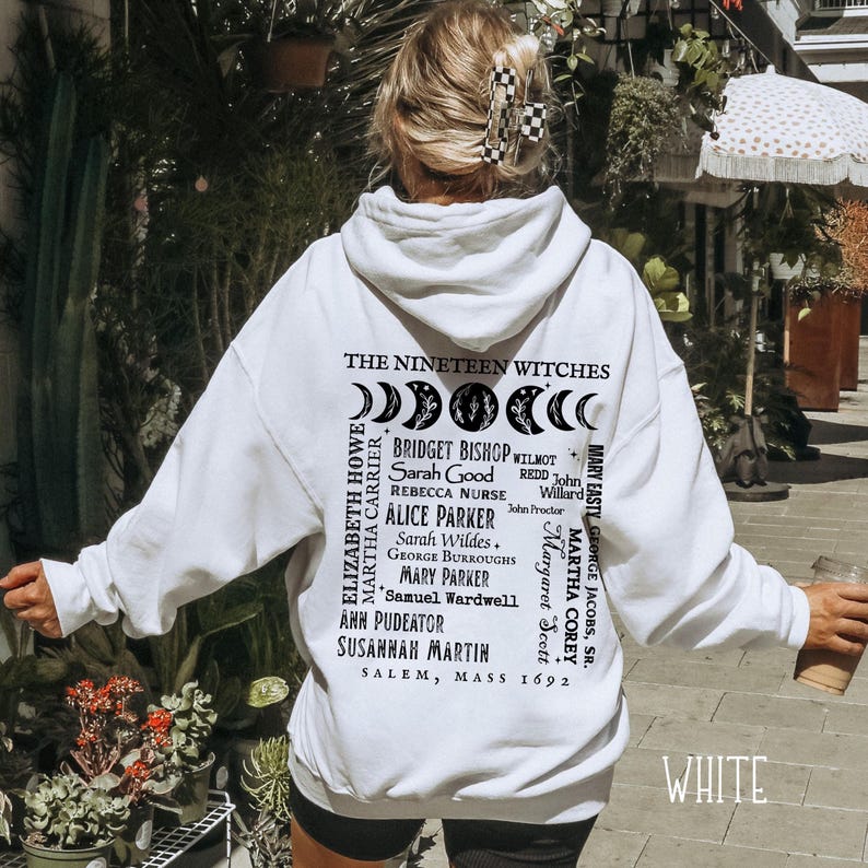 Op de afbeelding: Een witte hoodie met een zwarte afbeelding op de achterkant. Het ontwerp bevat maanfasen, de tekst THE NINETEEN WITCHES, een lijst met namen waaronder Bridget Bishop en Sarah Good, en de tekst SALEM, MASS 1692 in een zwart lettertype.