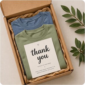Op de afbeelding: Een kartonnen verzenddoos met een opgevouwen blauw t-shirt en een opgevouwen olijfgroen t-shirt. Bovenop de shirts ligt een witte bedankkaart met de tekst thank you so much for your order, your support means the world to us, made with passion, just for you.