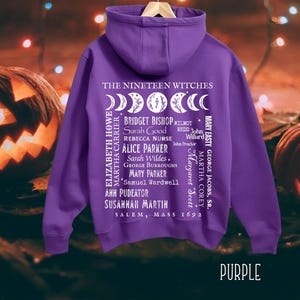 Op de afbeelding: Een paarse hoodie met een witte afbeelding op de achterkant. Het ontwerp bevat maanfasen, de tekst THE NINETEEN WITCHES, SALEM, MASS 1692 en een lijst met namen waaronder Bridget Bishop, Sarah Good, Rebecca Nurse en anderen.